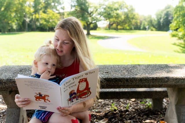 An au pair sitting on a park bench, reading a picture book to a toddler on her lap.
