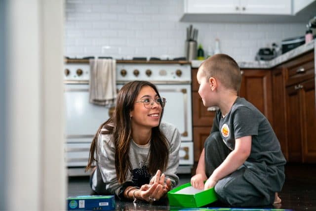An au pair sitting on the kitchen floor with a young boy, both smiling during a moment of play.